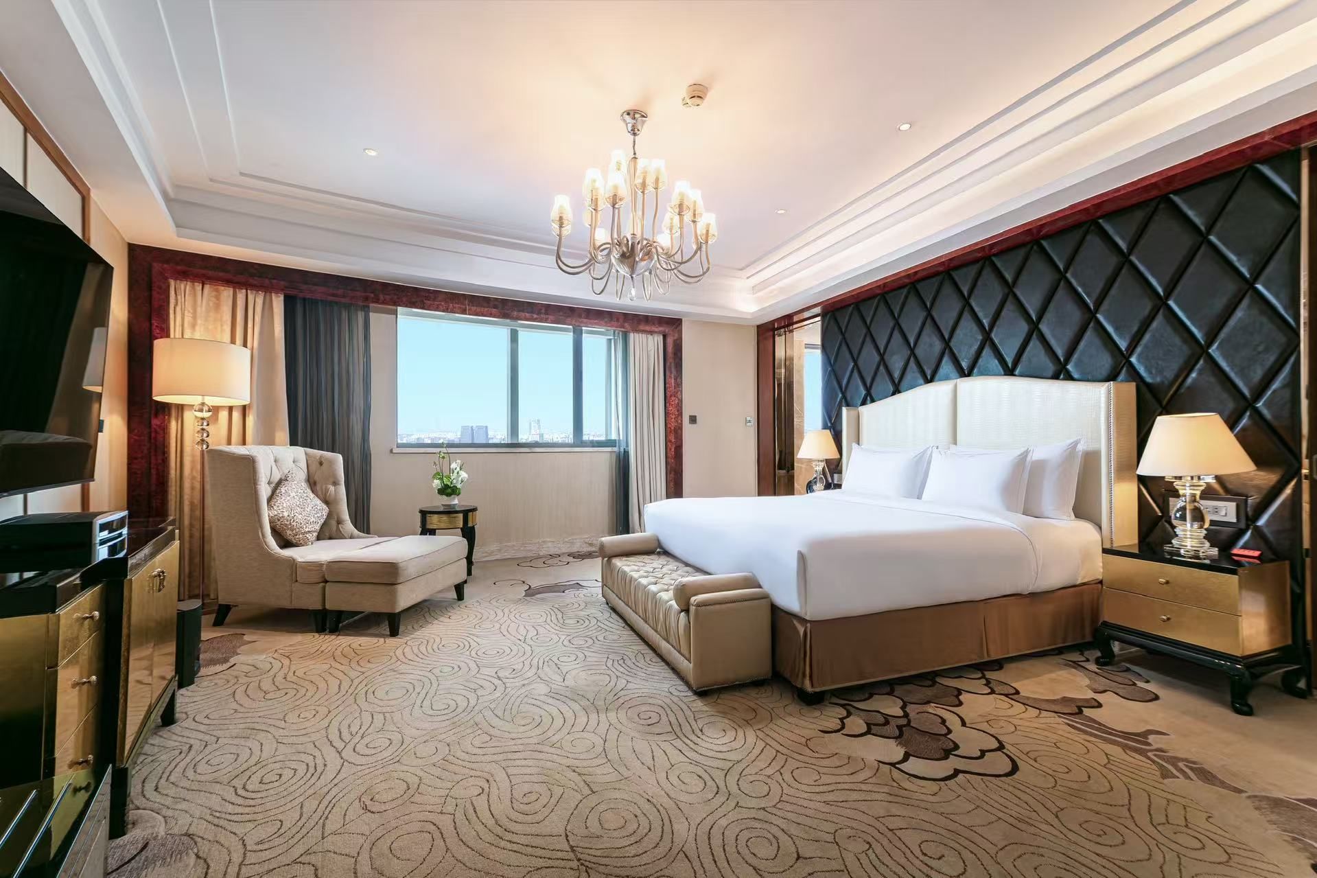 A hotel room with a large bed, a sitting area with a chair and a small table, a television, and a view of the cityscape.