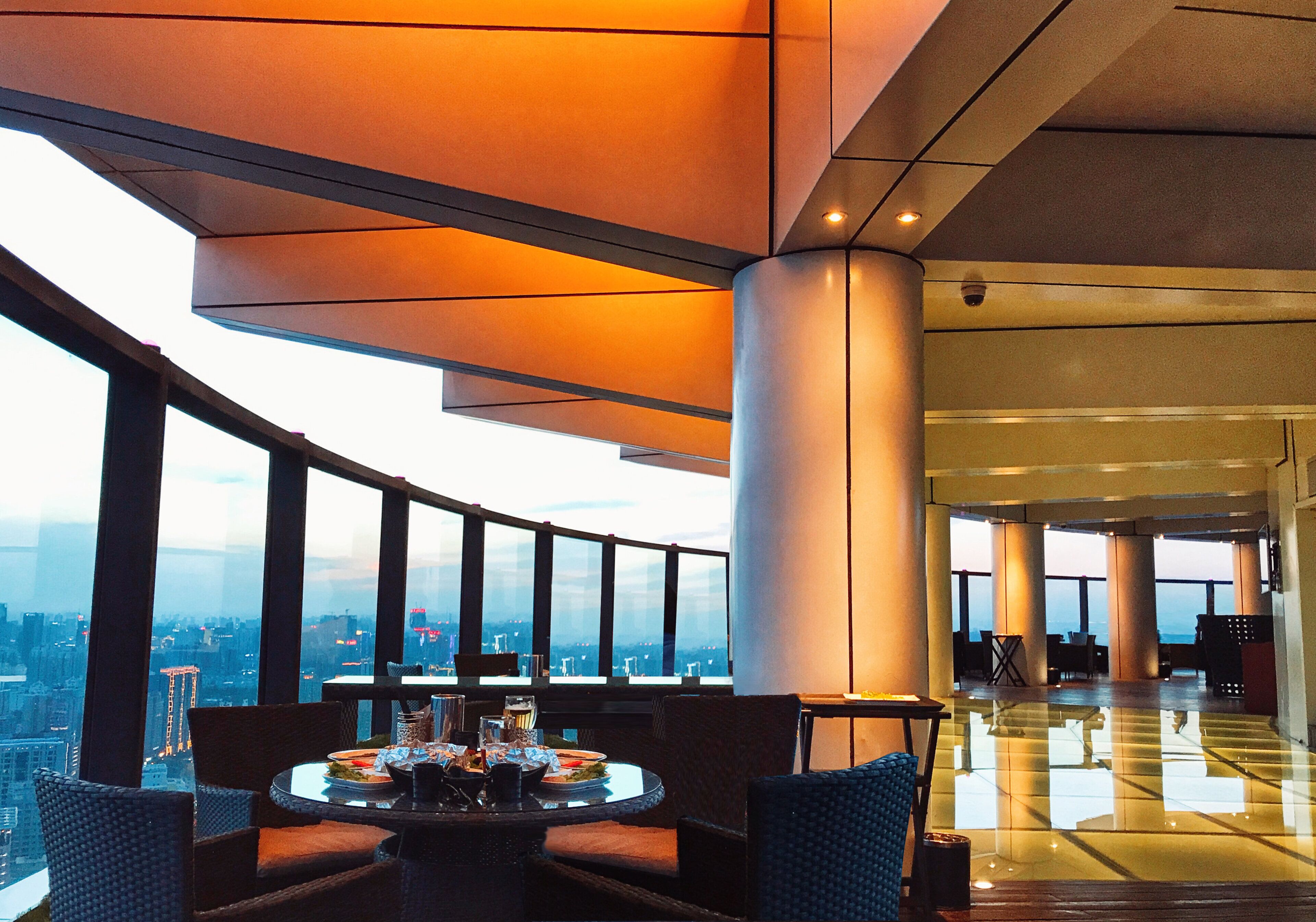 L’intérieur d’un hôtel de grande hauteur présente un espace de restauration avec des tables et des chaises rondes, de grandes fenêtres offrant une vue sur la ville et une architecture moderne caractérisée par de grandes colonnes.