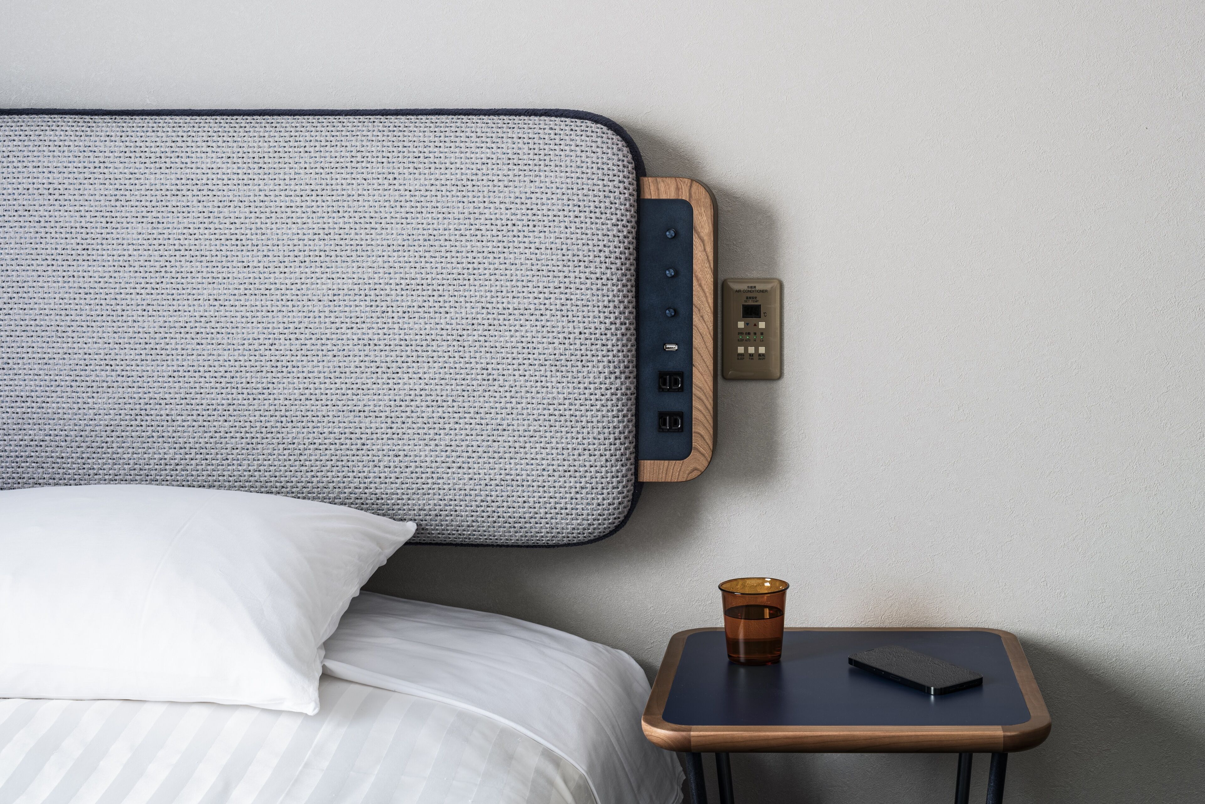 Una cama bien tendida con un almohadón blanco, una mesita de noche con una taza y un teléfono inteligente, y un enchufe de pared.