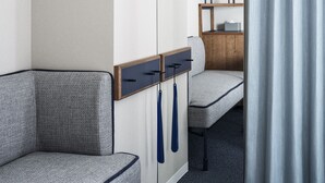 Una esquina de habitación de hotel con un sofá gris, un aparador de madera con estante y una cortina entreabierta.