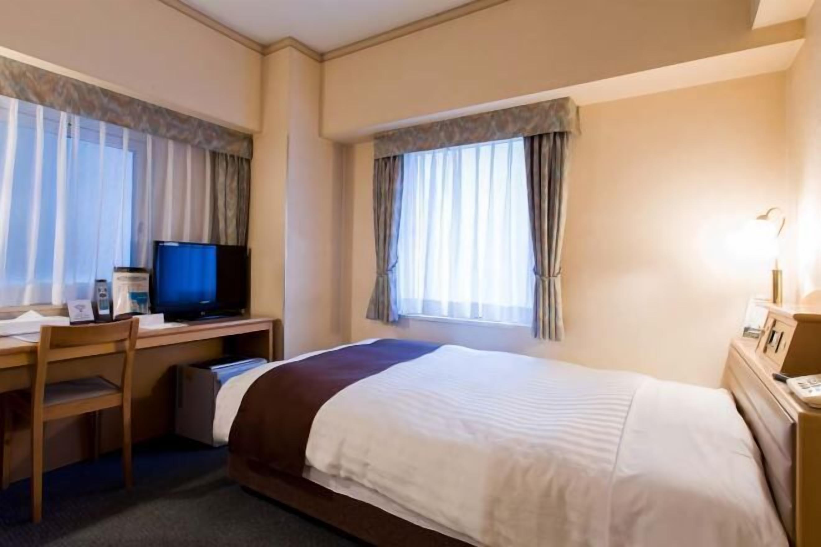 Habitació d'hotel amb un llit, una taula de nit amb una làmpada, un escriptori amb un ordinador i una televisió.