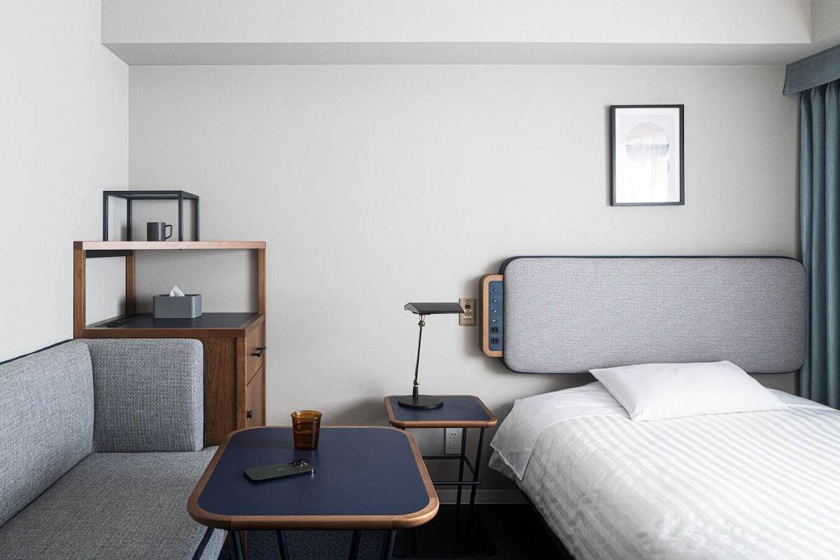 Une chambre d’hôtel avec un lit, un bureau, une chaise, une petite table, une lampe, une étagère et un tableau au mur.