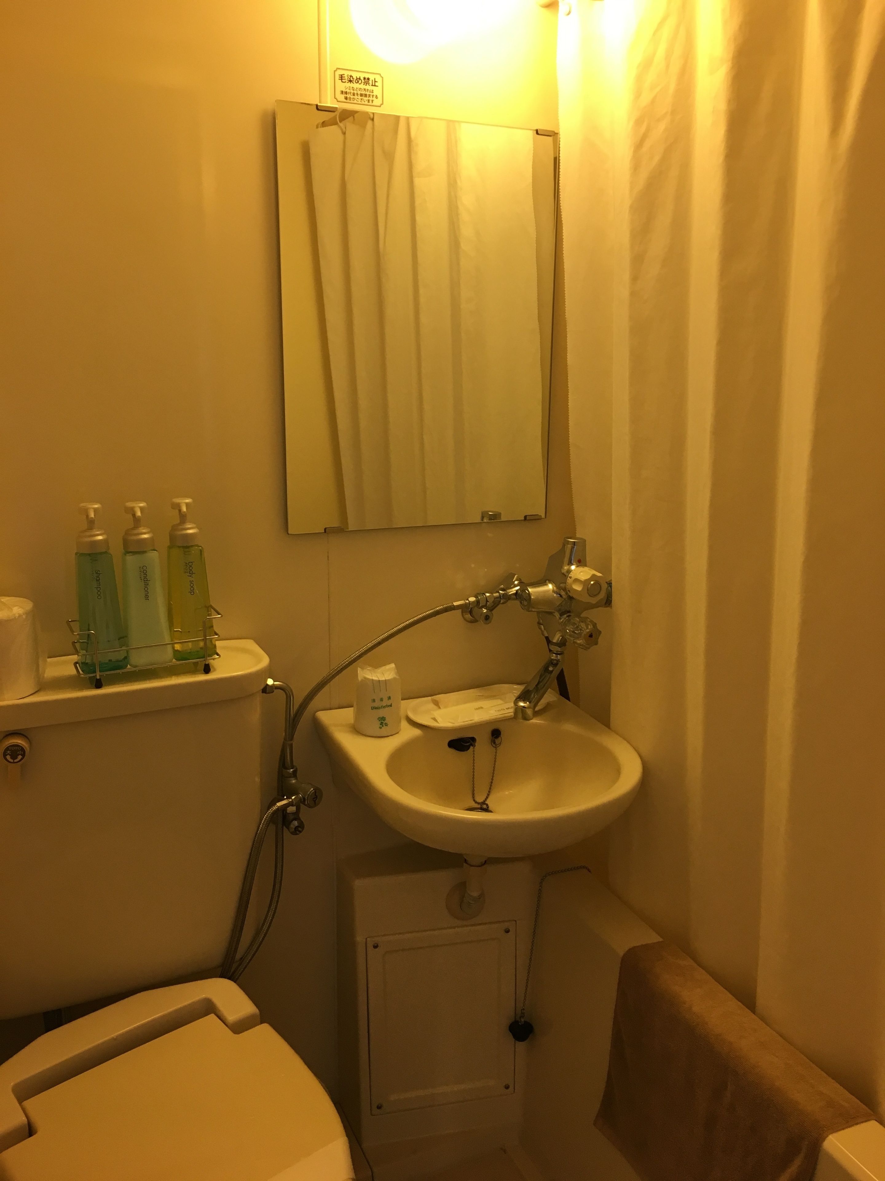 A bathroom with a toilet, sink, and mirror. There are three bottles on the shelf above the toilet.