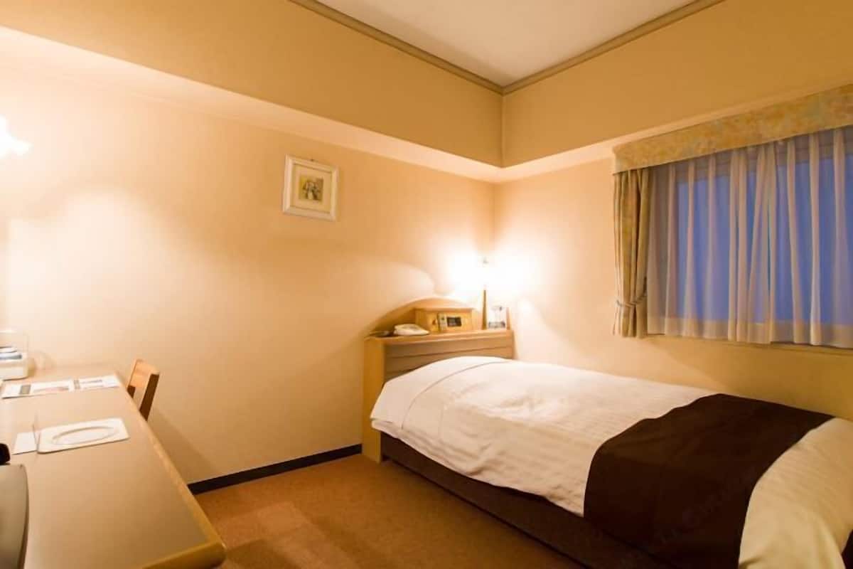 Une chambre d’hôtel avec un lit simple, une table de chevet avec une lampe, une petite table avec une chaise et un tableau encadré au mur.