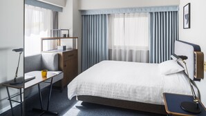 Una habitación de hotel moderna con una cama bien hecha, un escritorio con una silla, una mesita pequeña y una lámpara de noche.