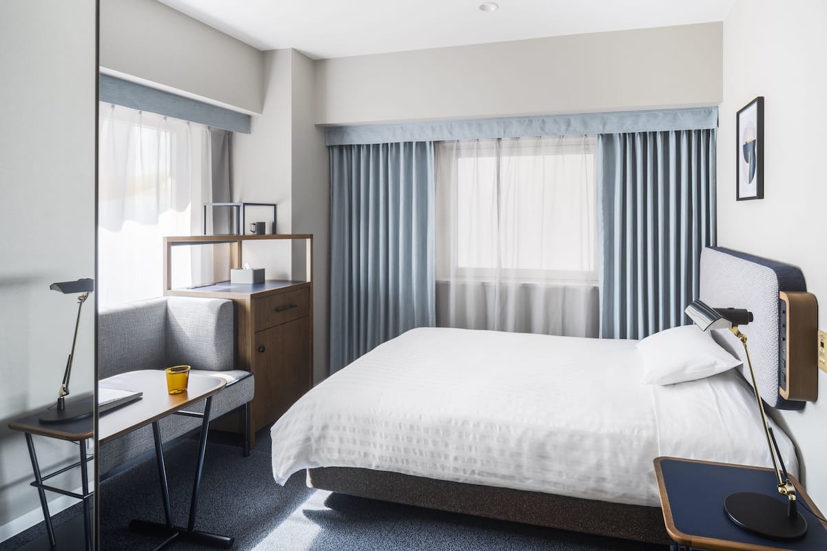 Une chambre d’hôtel moderne avec un lit soigneusement fait, un bureau avec une chaise, une petite table et une lampe de chevet.
