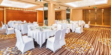 Un salón de banquetes con mesas redondas dispuestas para comer, sillas blancas y suelo alfombrado.