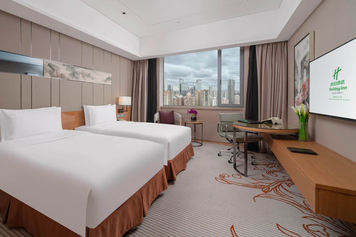 Habitación de hotel con dos camas, un escritorio, una silla, una mesita, una lámpara, un jarrón con flores y vistas a la ciudad desde la ventana.