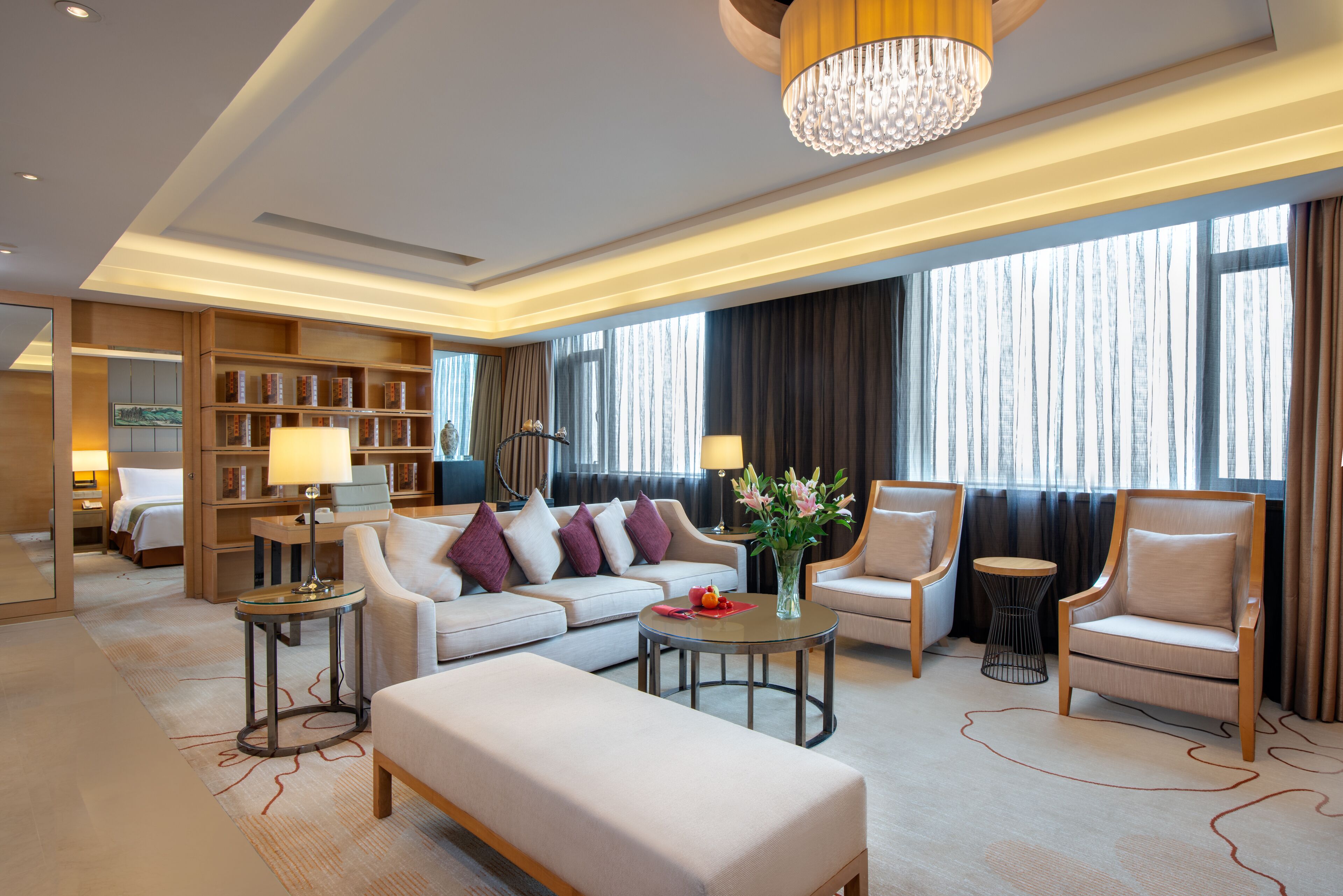 A modern hotel lobby with a sofa, armchairs, a round coffee table, and a bookshelf.