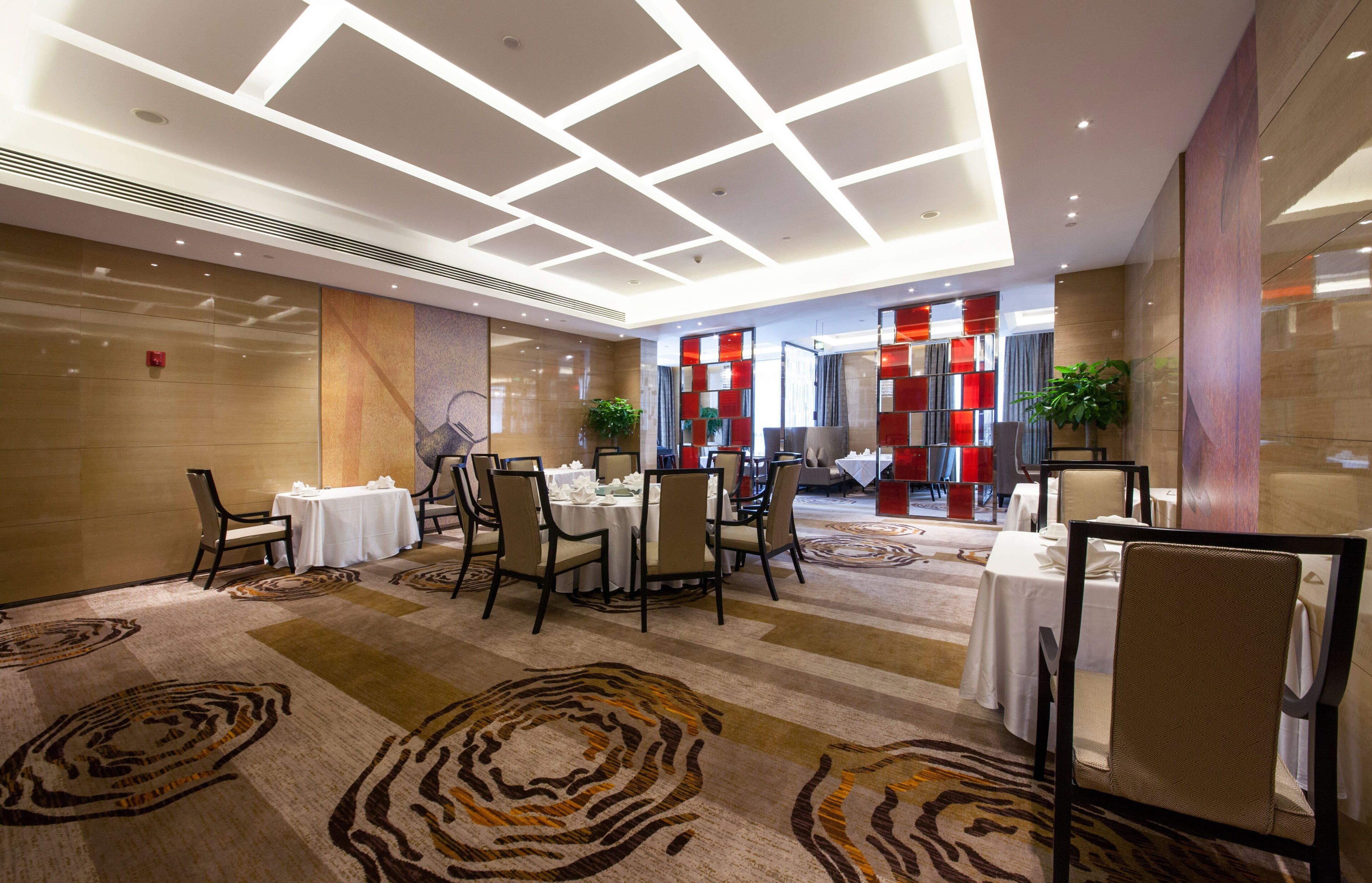 Un comedor de hotel con mesas dispuestas para los huéspedes, una alfombra estampada y un diseño de techo moderno.