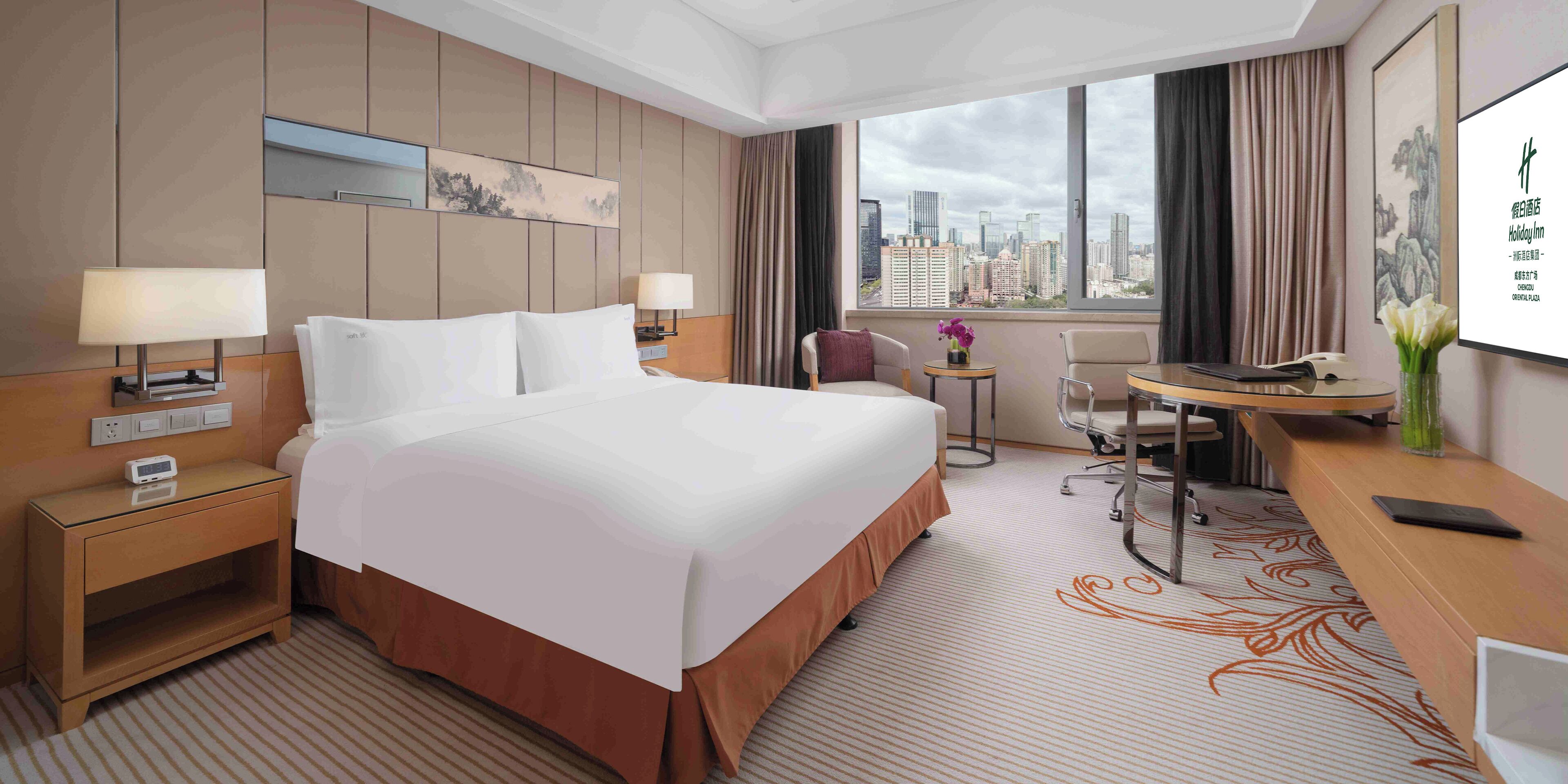 Quarto de hotel com uma cama grande, mesas de cabeceira, uma escrivaninha, uma cadeira e vista para a paisagem urbana.