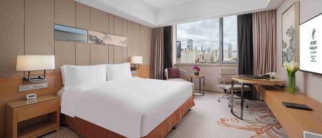 A hotel room with a large bed, bedside tables, a desk, a chair, and a view of the cityscape.