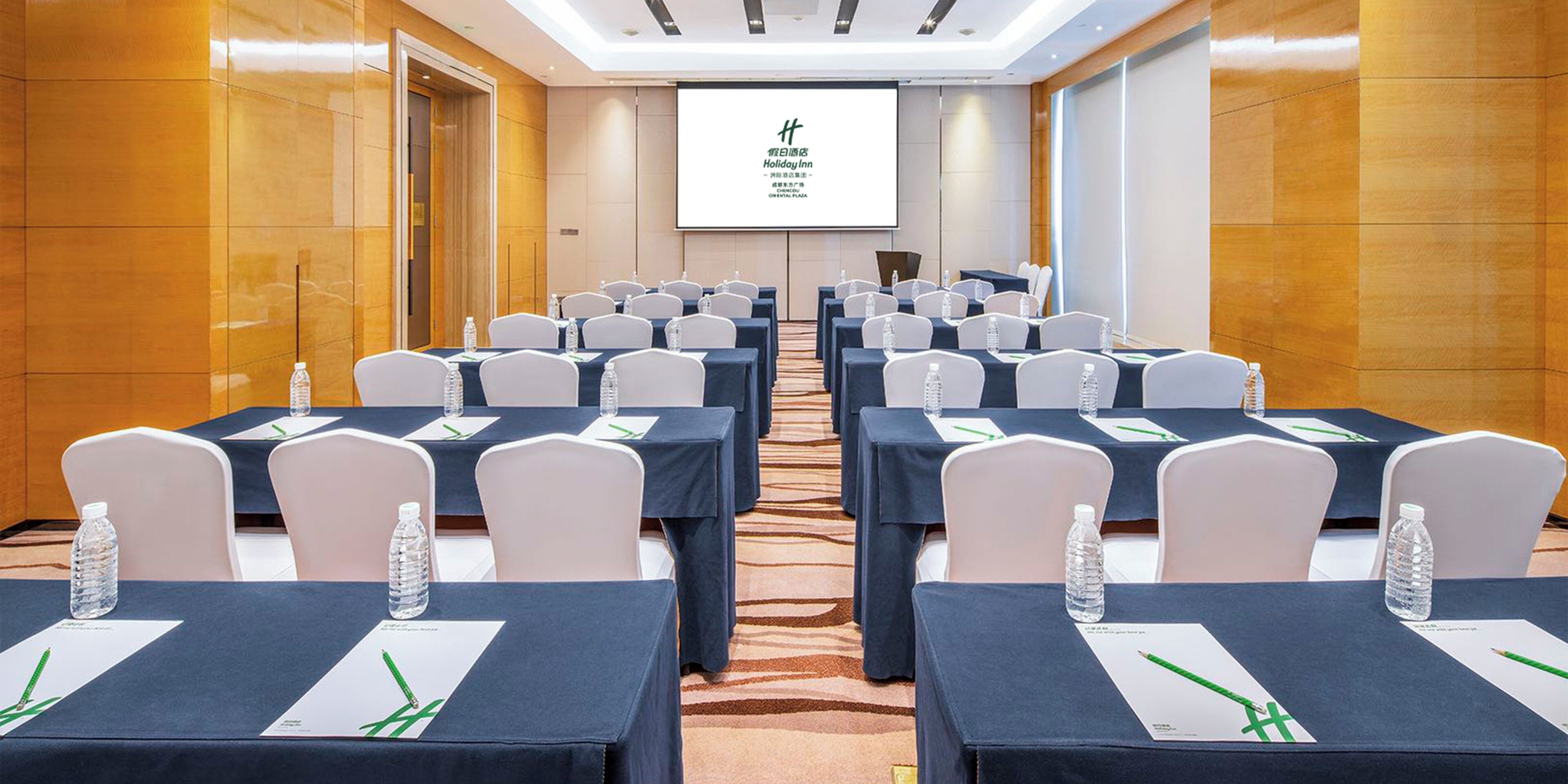 A conference room with rows of tables, chairs, and a projection screen displaying the hotel name.