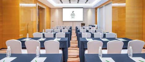A conference room with rows of tables, chairs, and a projection screen displaying the hotel name.