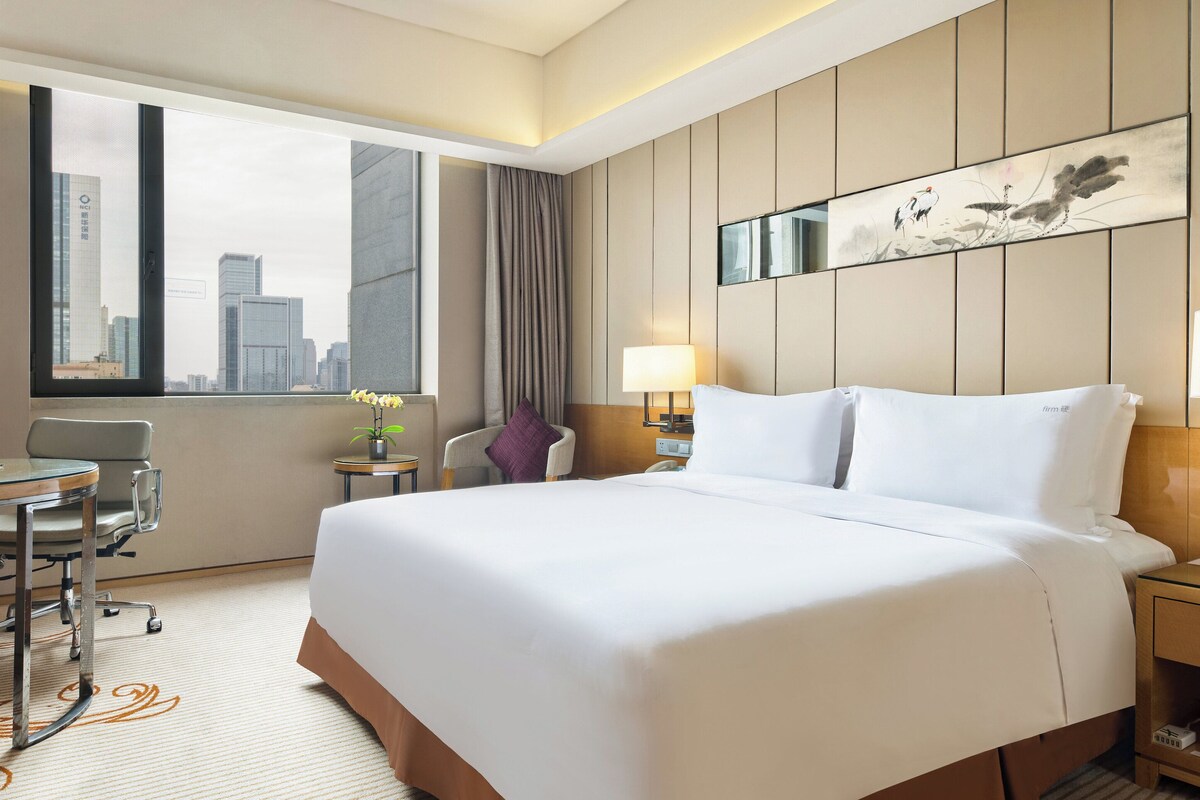 Habitación de hotel con una cama grande, un escritorio con una silla, una mesita con una lámpara y vistas a la silueta urbana a través de la ventana.
