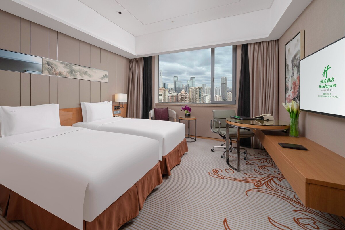 A hotel room with two beds, a desk, a chair, a small table with a vase, and a city view through the window.