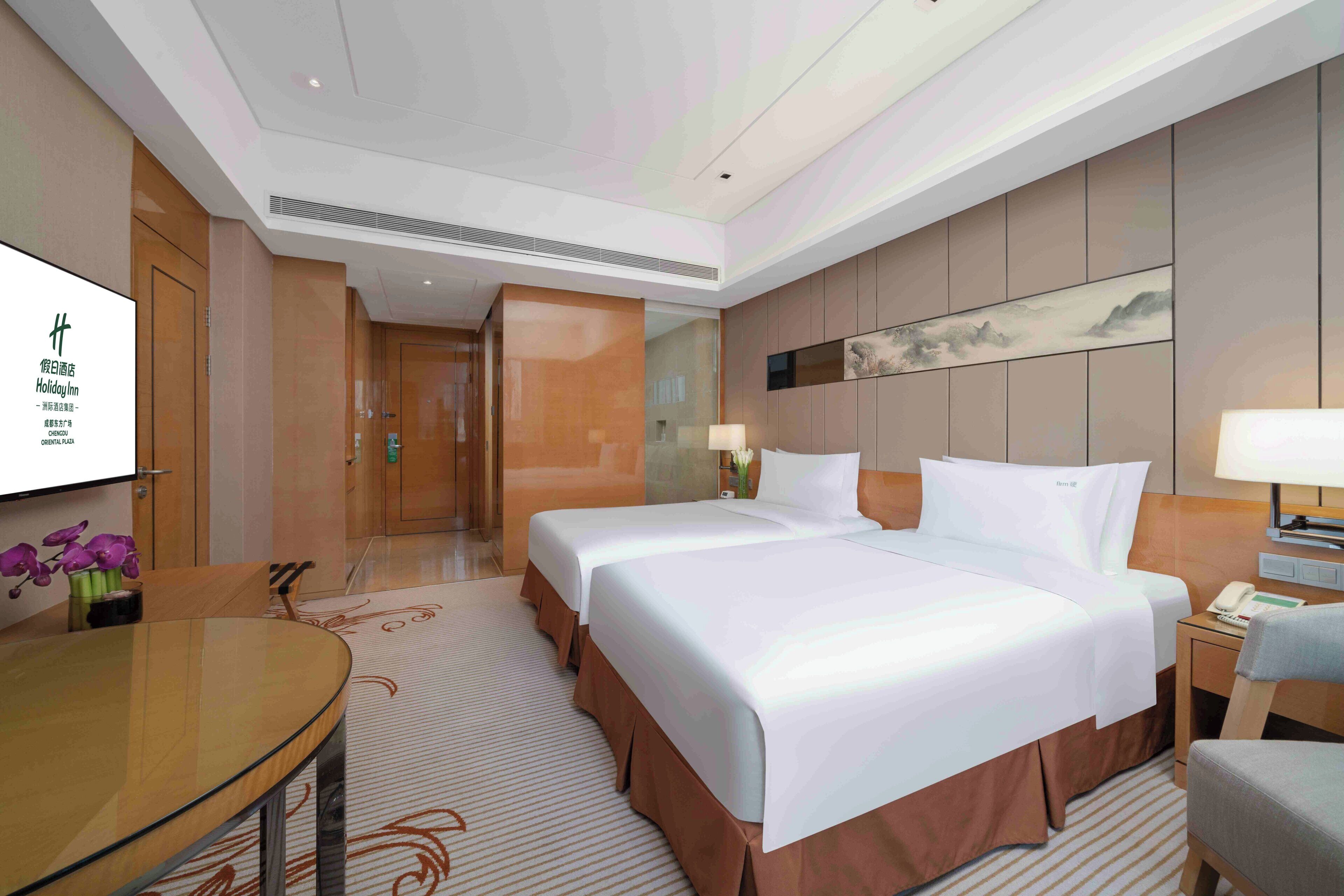 A hotel room with two beds, a desk, a chair, a TV, and a painting on the wall.