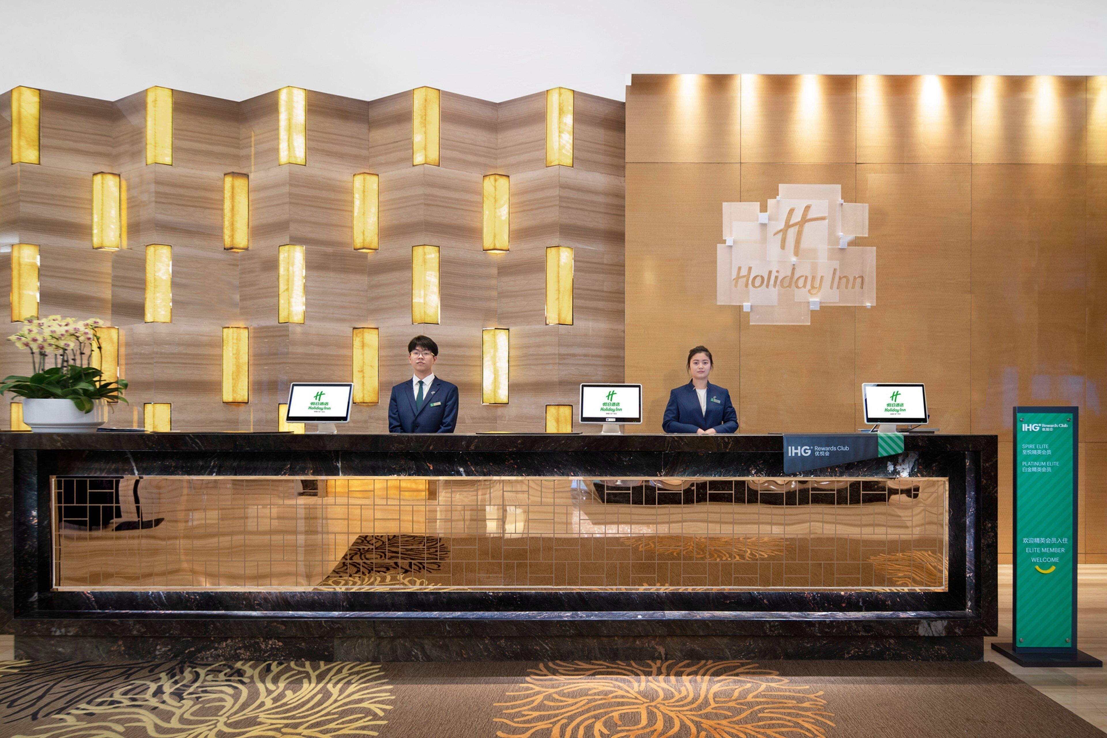 A reception desk with two staff members, a wooden wall with vertical light fixtures, and a sign for Holiday Inn.