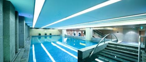 An indoor swimming pool with clear water, surrounded by stone walls and a staircase leading to an upper level.