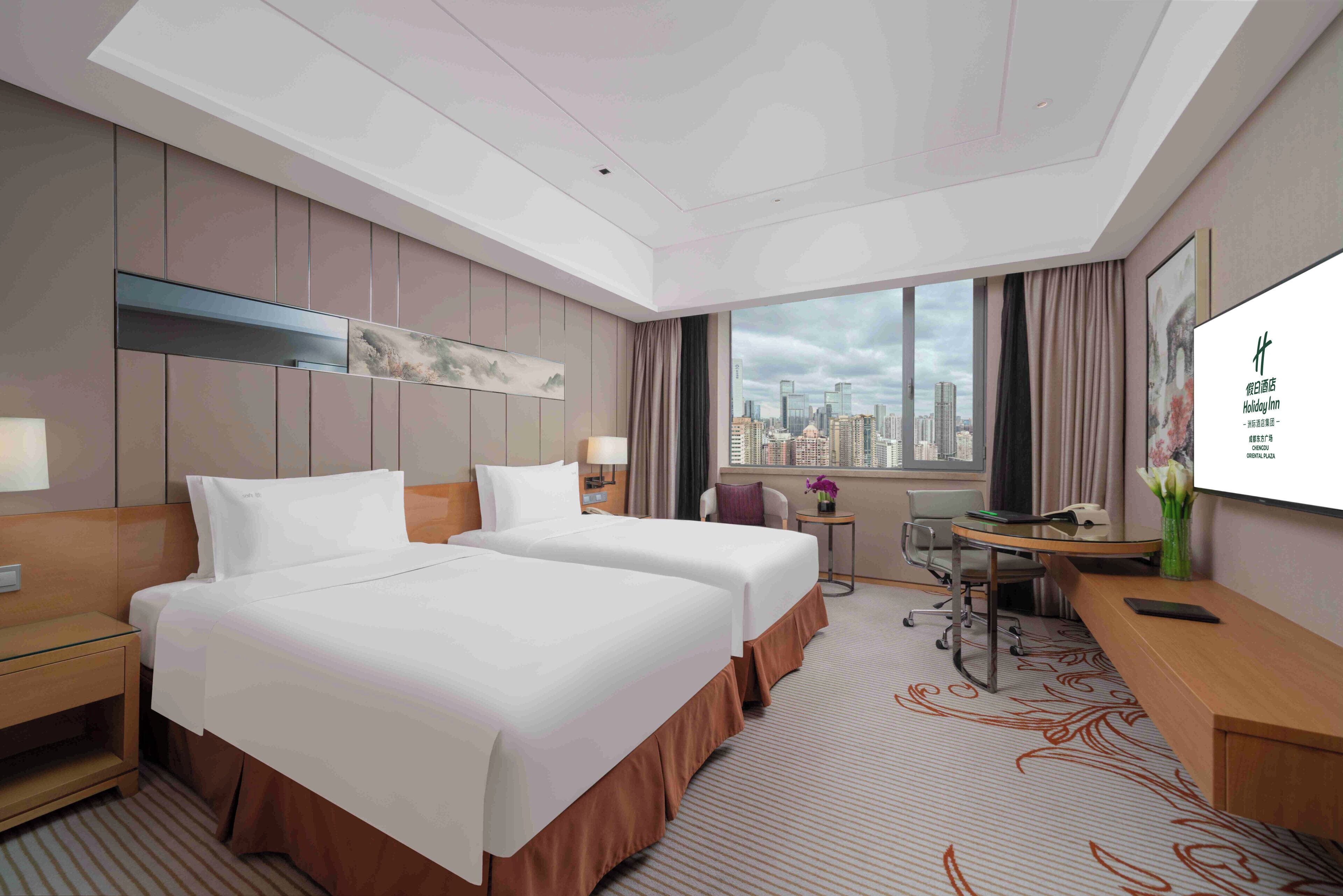 A hotel room with two beds, a desk, a chair, a TV, and a window with a city view.