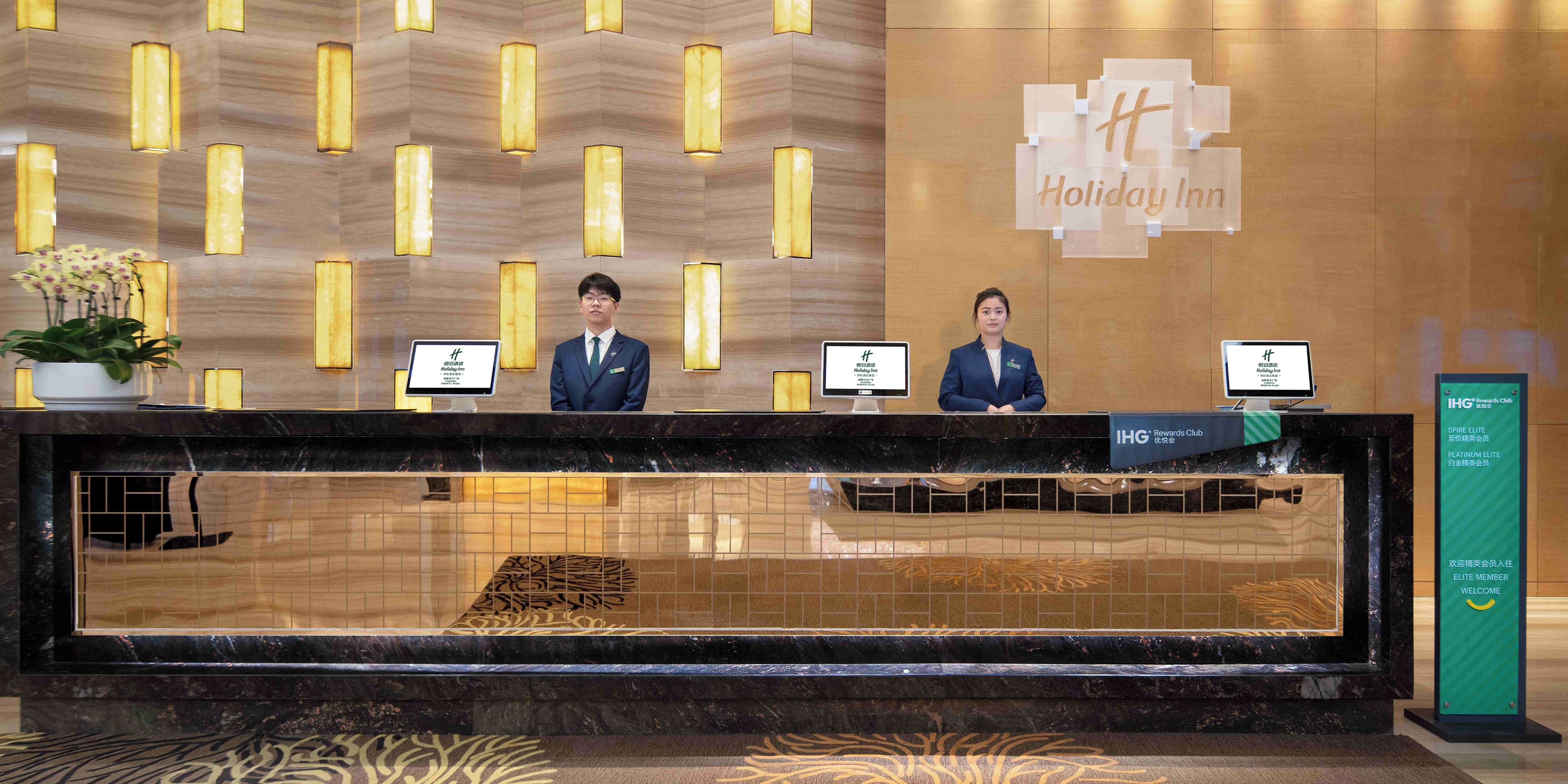 A hotel reception desk with two staff members, a sign for the Holiday Inn, and a sign for the IHG Rewards Club.