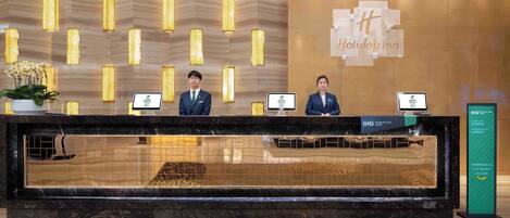 A hotel reception desk with two staff members, a sign for the Holiday Inn, and a sign for the IHG Rewards Club.
