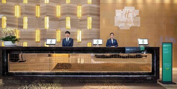A hotel reception desk with two staff members, a sign for the Holiday Inn, and a sign for the IHG Rewards Club.
