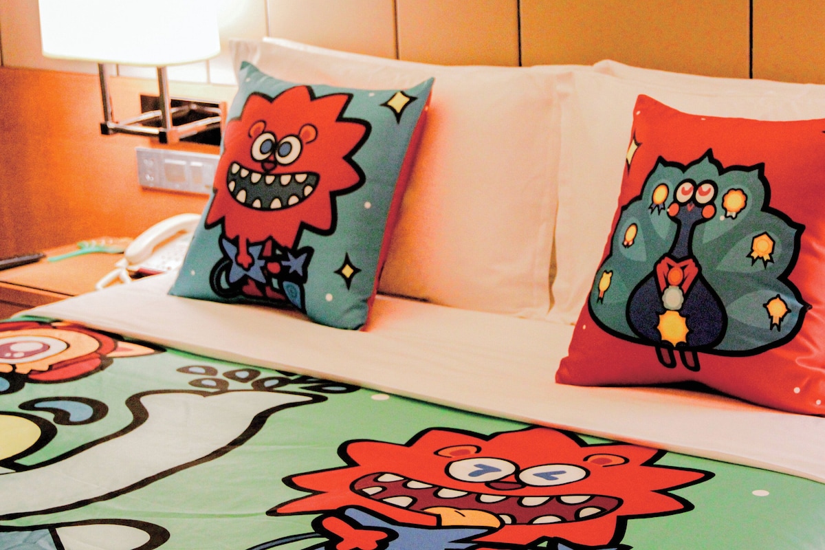 A hotel room with two pillows featuring cartoon characters, a bed with a colorful bedspread, a bedside table with a lamp, and a nightstand with a phone and remote control.