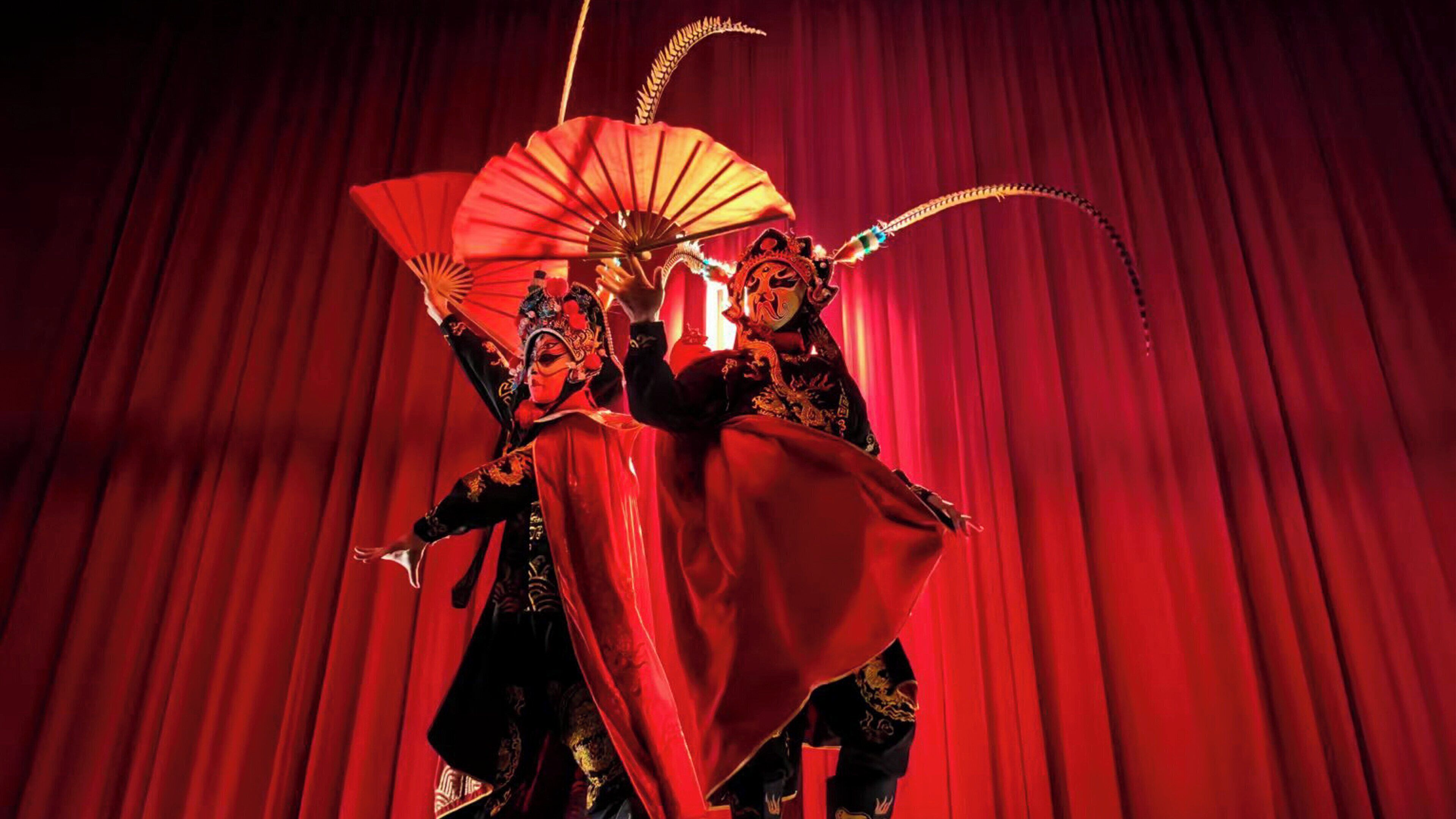 Two performers in traditional costumes with a red curtain backdrop.
