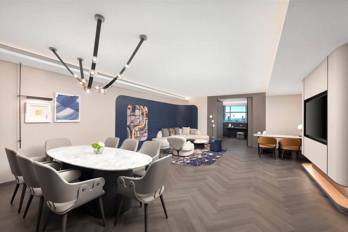 A modern hotel room with a round dining table, comfortable seating, a large TV, and a view of the city.