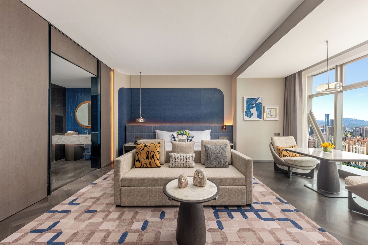 A modern hotel room with a large bed, a sofa, a dining table, and a city view.