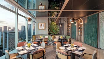 A high-rise restaurant with multiple dining tables and booths, large windows offering city views, and a green wall with plants.