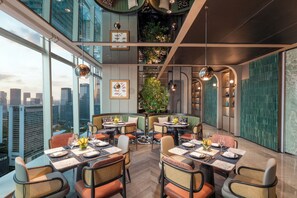 Un restaurante de altura con múltiples mesas y cabinas, amplios ventanales con vistas a la ciudad y una pared verde con plantas.