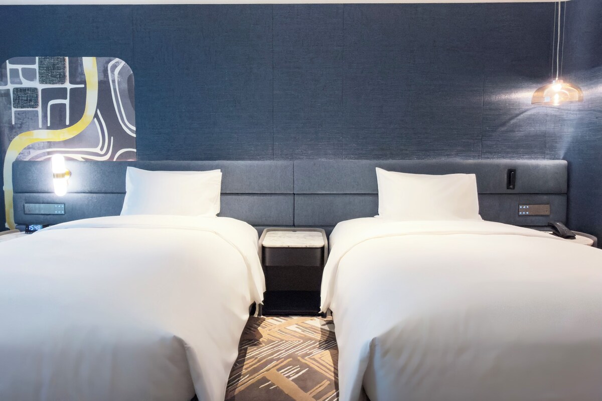 A hotel room with two beds, a nightstand, a digital clock, and a wall with a geometric design.
