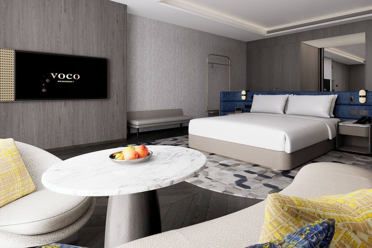 Ein modernes Hotelzimmer mit Bett, Sitzgelegenheit und einem Fernseher, der 'Voco' anzeigt.