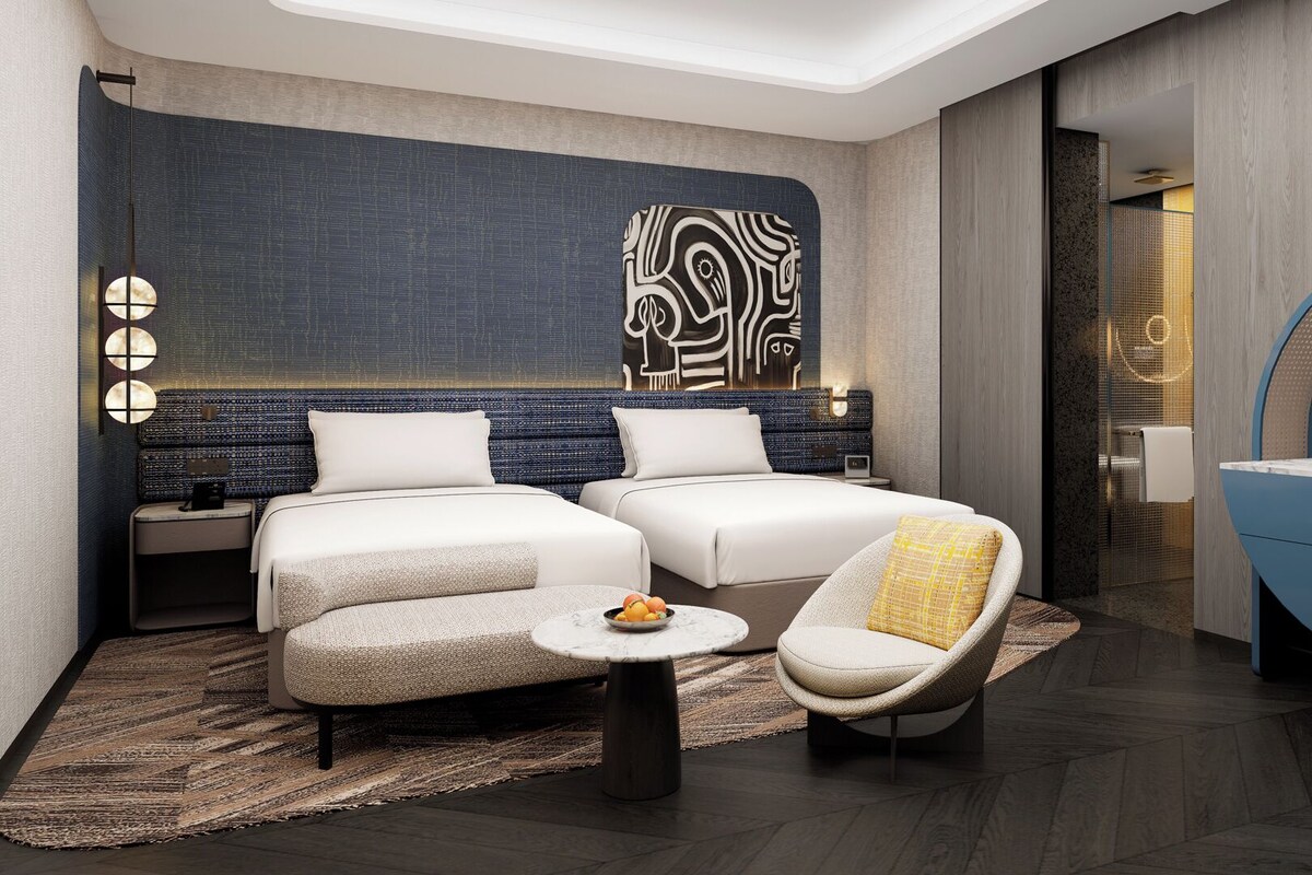 A modern hotel room with two beds, a round coffee table, a chair, and a blue accent wall.