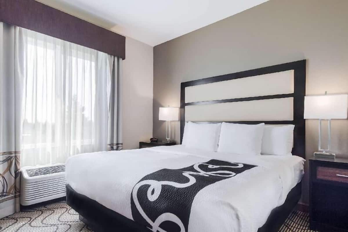 Ein Hotelzimmer mit einem großen Bett, zwei Nachttischlampen, einem Heizgerät und einem Fenster mit durchsichtigen Vorhängen.