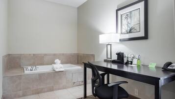 A bathroom with a bathtub, a toilet, a sink, a desk with a chair, a framed picture on the wall, and a lamp.
