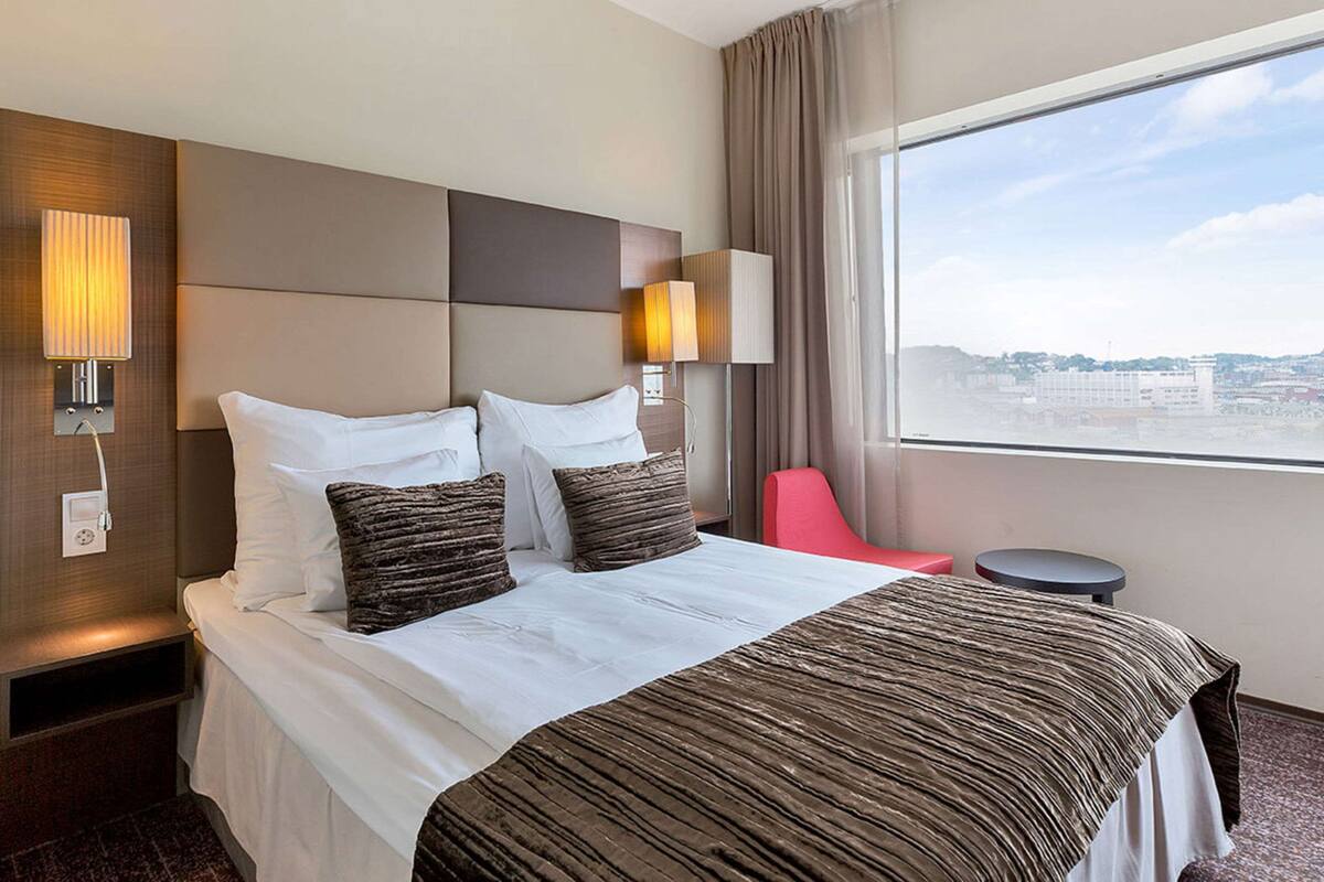 A hotel room with a large bed, two bedside lamps, a red chair, a small table, and a window with a city view.