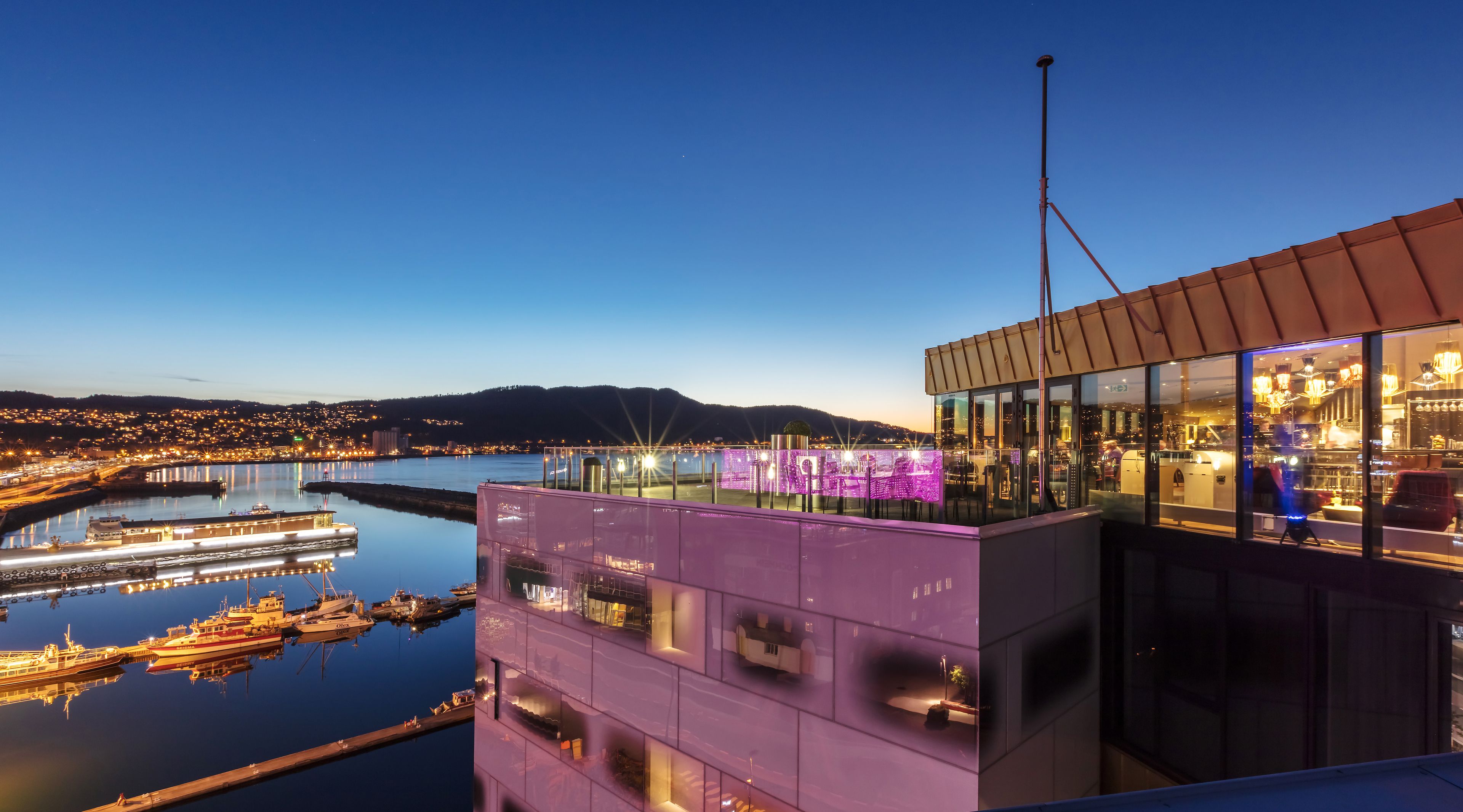 Um edifício moderno com fachada de vidro, com vista para um rio, apresentando um cais com barcos e um horizonte urbano ao fundo.
