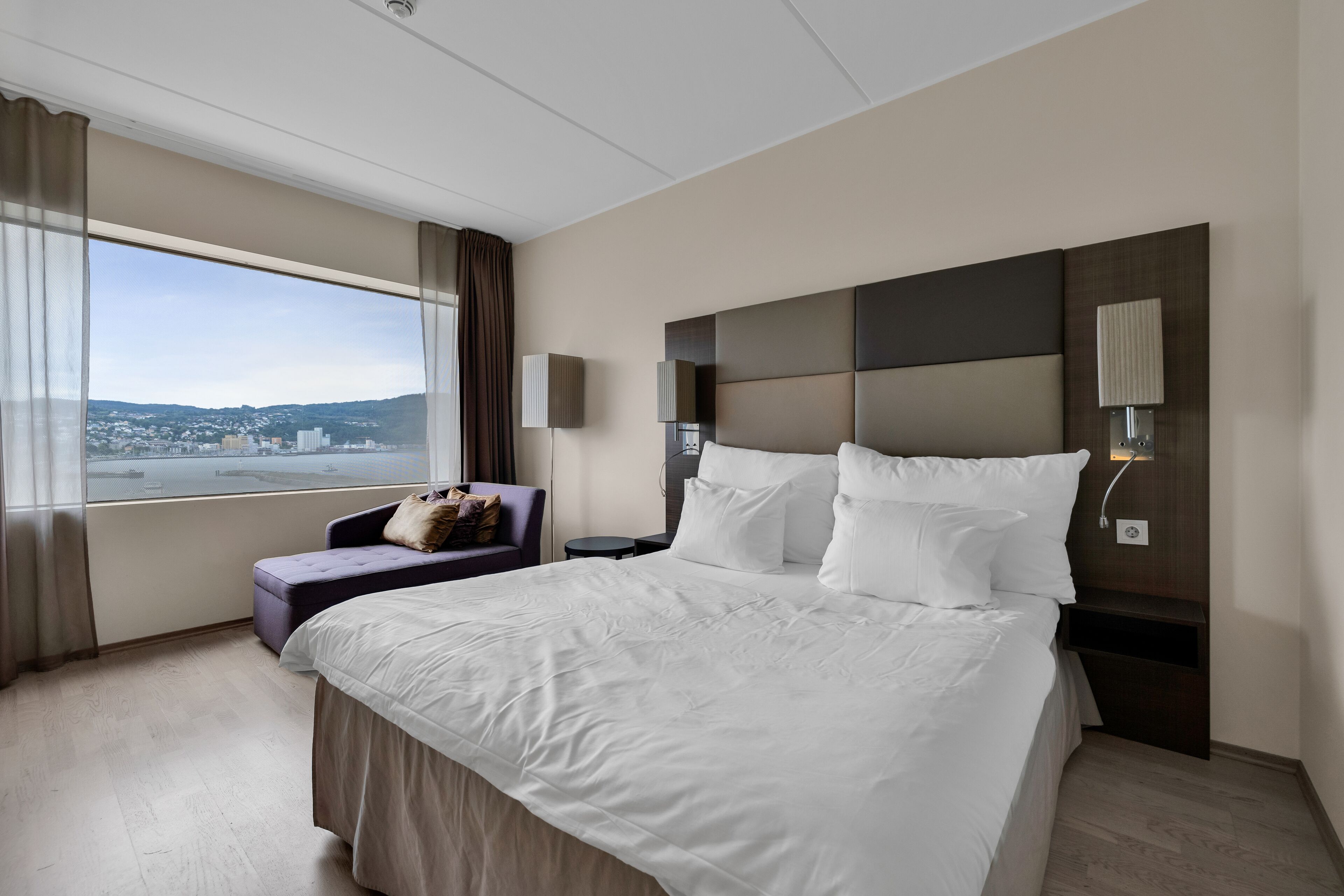Habitación de hotel con una cama grande, vistas a la ciudad, un sofá y una mesita de noche con una lámpara.