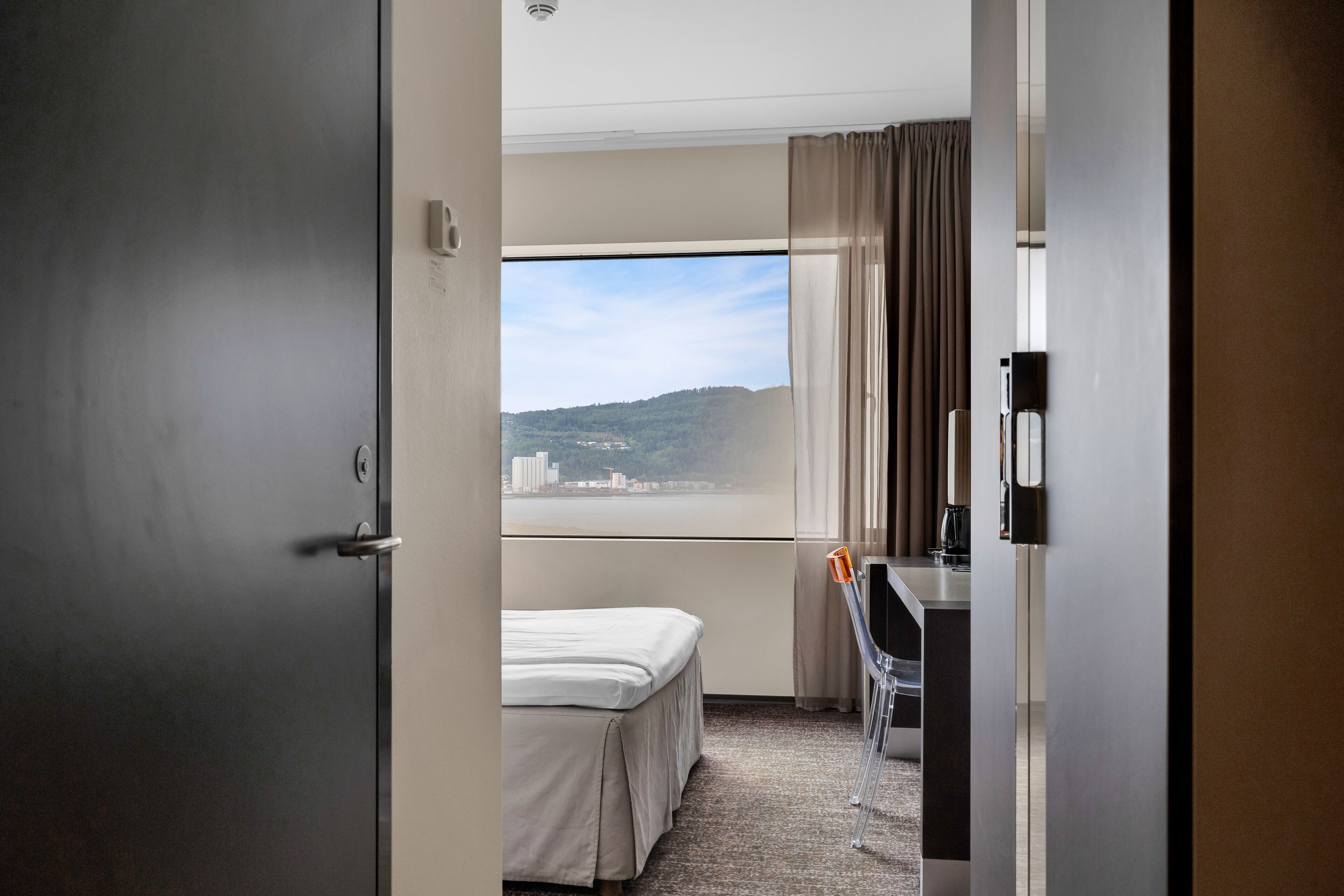 A hotel room with a bed, a desk, and a view of a cityscape through a large window.