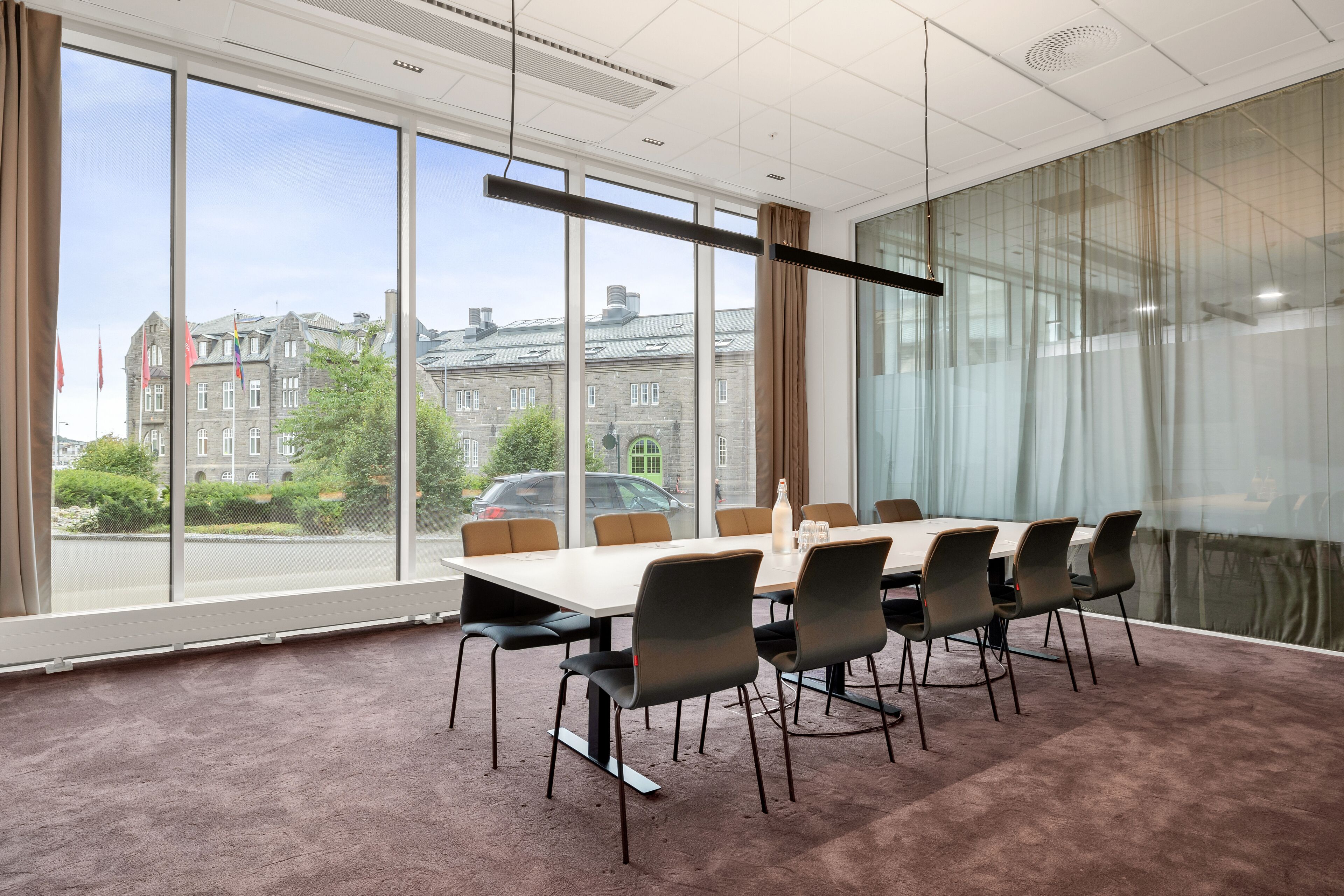 A conference room with a large table and chairs, large windows with a view of buildings and trees, and a ceiling with recessed lighting.