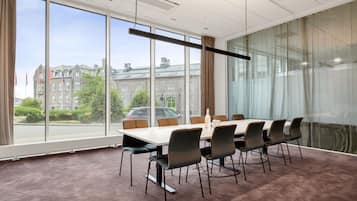 A conference room with a large table and chairs, large windows with a view of buildings and trees, and a ceiling with recessed lighting.