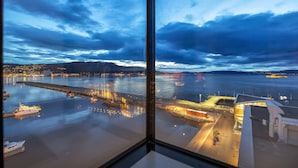 Une vue sur le front de mer, depuis une fenêtre donnant sur une marina avec des bateaux, un quai et des bâtiments dont les fenêtres sont illuminées.