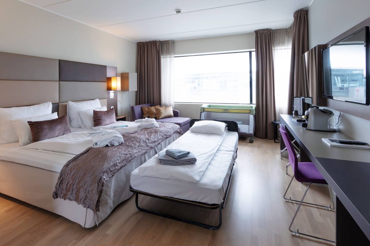 A hotel room with a large bed, a smaller bed, a desk, a chair, a TV, and a window with curtains.