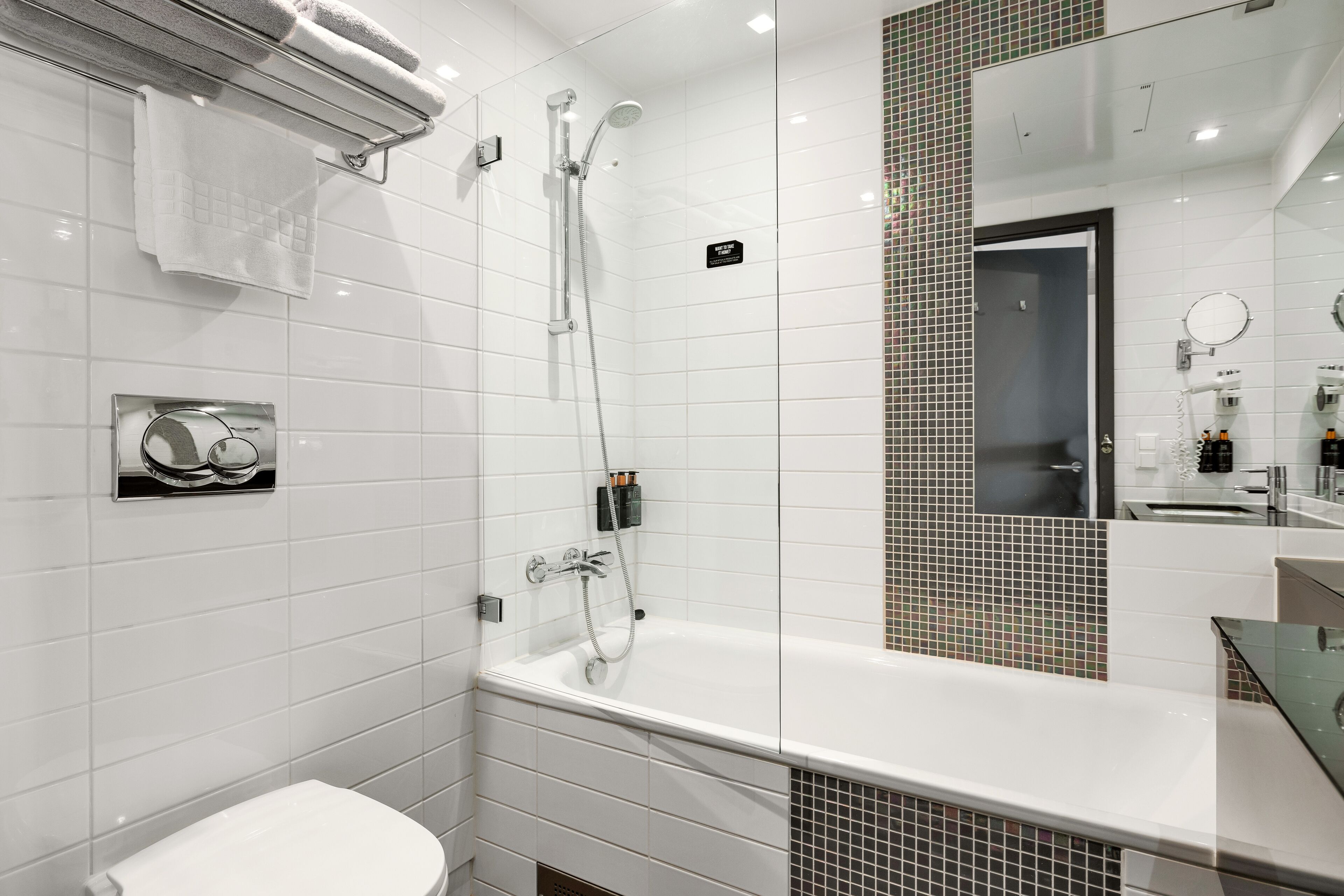 A modern bathroom with a white tiled wall, a glass shower enclosure, a white toilet, and a large mirror with a chrome frame.