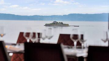A dining table with white tablecloths and wine glasses, overlooking a calm lake with a small island and mountains in the background.