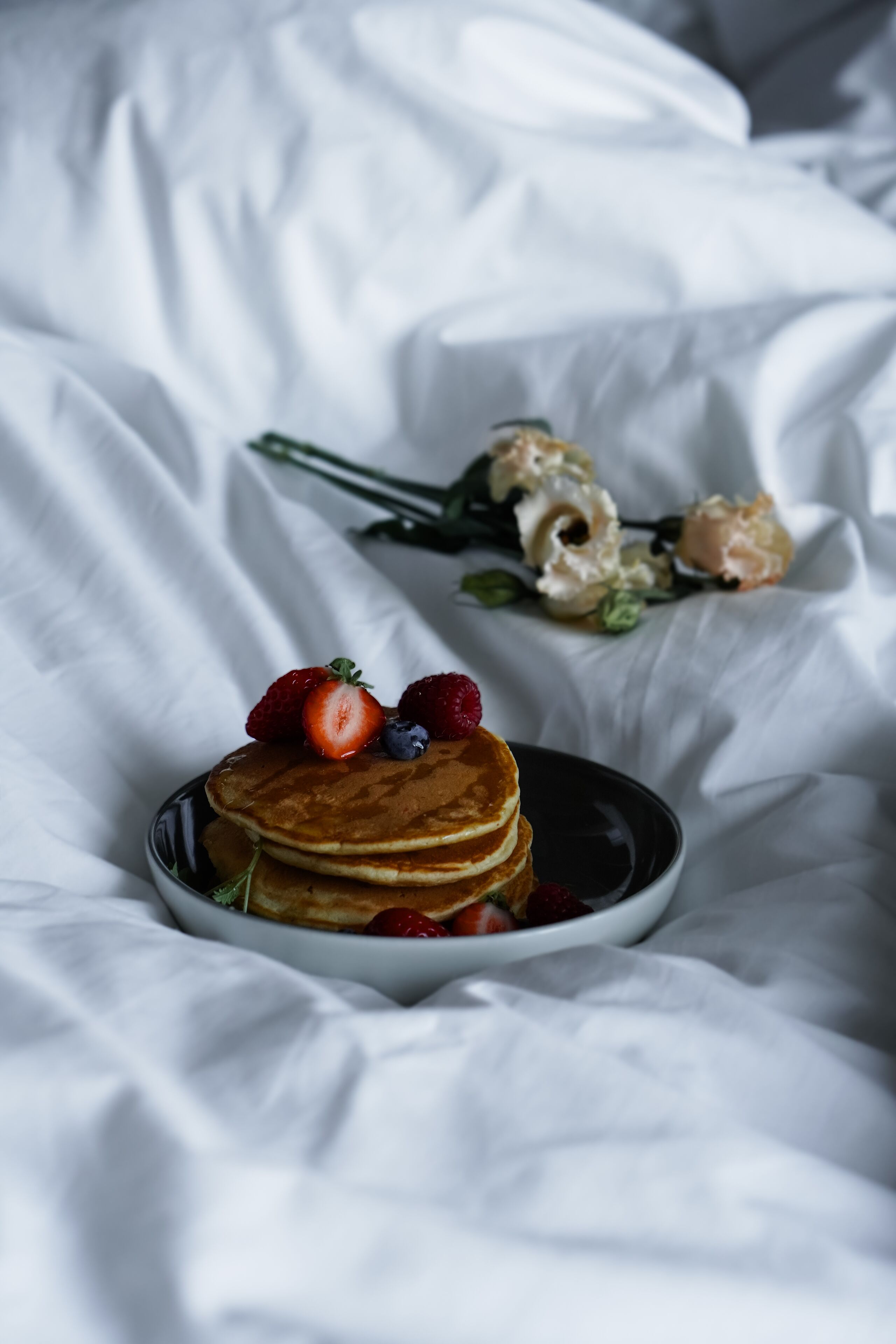Ein Stapel Pfannkuchen auf einem Teller mit Erdbeeren und Blaubeeren obenauf, auf einer weißen Bettdecke platziert.