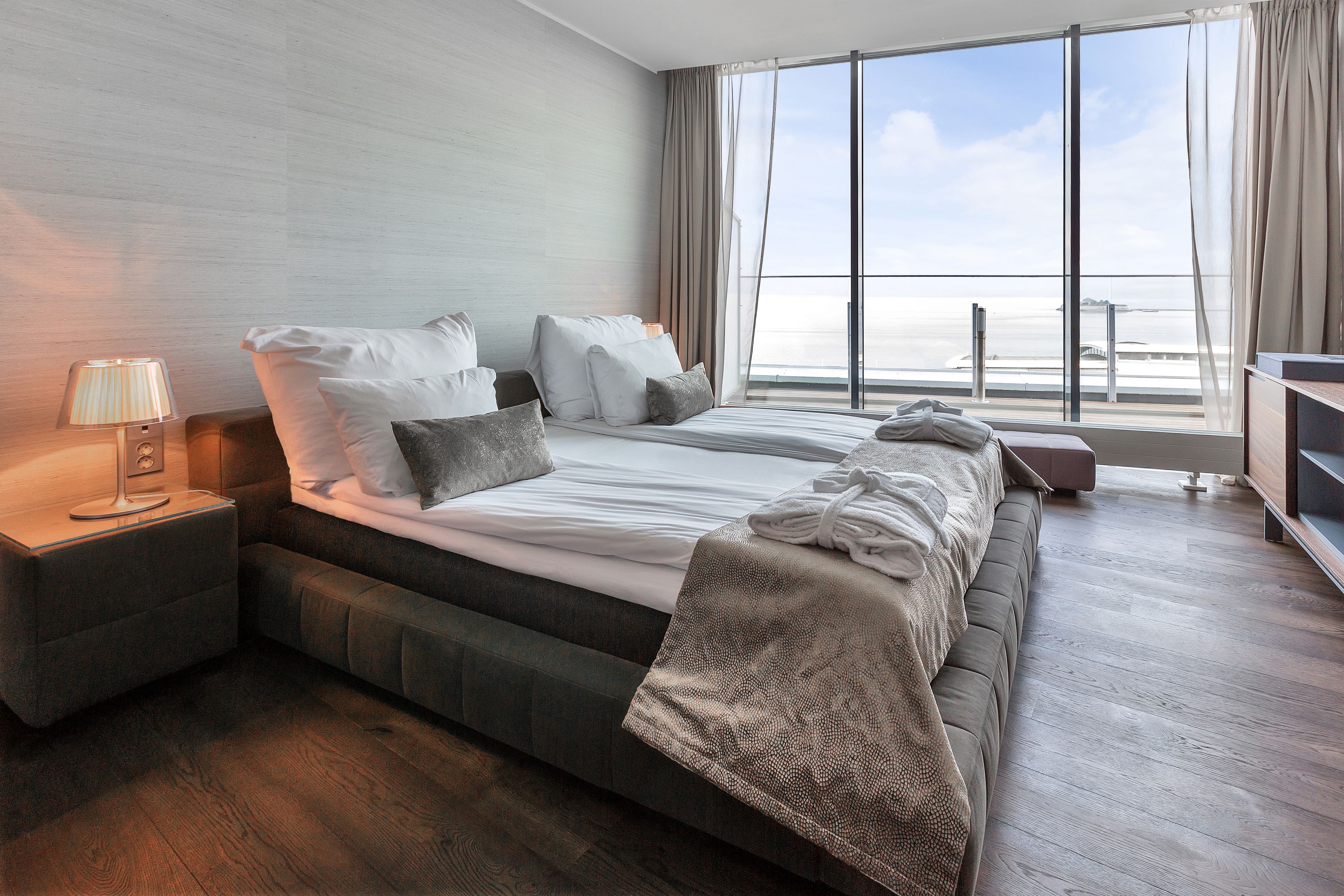 A modern hotel room with a large bed, a bedside table with a lamp, a TV, and a view of the sea through large windows.